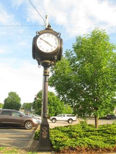 a clock on a pole in the middle of a parking lot with cars parked behind it