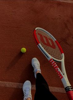 a tennis ball and racket on a court with someone's feet in the foreground