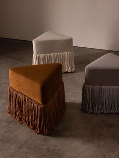 three foot stools with fringed covers on them in an empty room, one is white and the other is brown