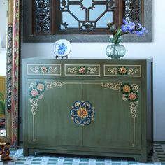 a green cabinet with flowers on it in front of an ornate mirror and vases