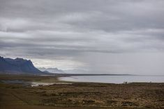 two horses are grazing on the grass by the water in front of some mountains and clouds