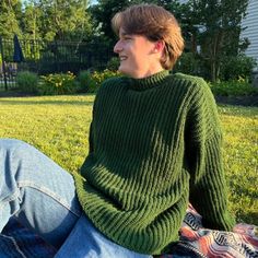 a woman sitting in the grass wearing a green knitted turtle neck sweater and jeans