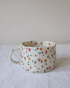 a white cup with colorful flowers on it sitting on top of a bed next to a wall