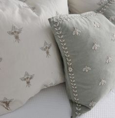 two pillows on top of a bed with white linens and silver embroidered bee designs