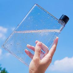 a hand holding a water bottle in front of a blue sky with clouds and trees