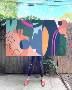 a woman standing in front of a wooden fence holding up a large piece of art