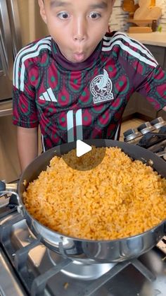 a young boy standing in front of an oven holding a pan filled with rice and looking at the camera