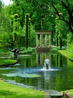 a pond in the middle of a lush green park with statues and trees around it