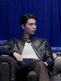 a man sitting on top of a couch in front of a blue curtain wearing a leather jacket