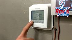 a person points at an electronic thermostaer on a wall that reads rox's