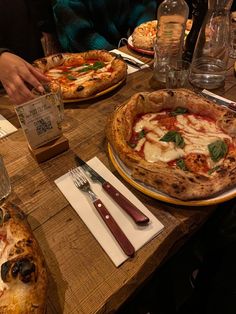 two pizzas sitting on top of a wooden table next to silverware and glasses