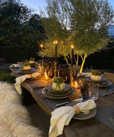 an outdoor table set for dinner with candles and plates on it, surrounded by greenery