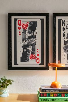 two framed pictures hang on the wall above a table with books and an orange lamp