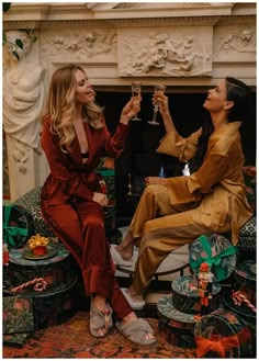 two women sitting next to each other in front of a fire place holding wine glasses