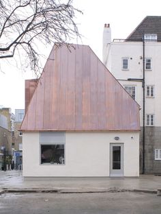 a small white building with a red roof in the middle of an empty lot next to some buildings