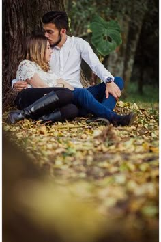 a man and woman sitting next to each other under a tree in the woods with leaves on the ground