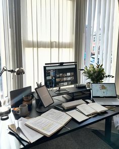 a desk with a computer, laptop and books on it in front of a window