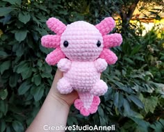 a pink crocheted stuffed animal is held by someone's hand in front of some bushes