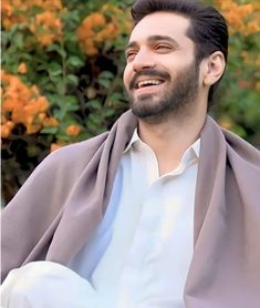 a man with a goatee smiles while sitting in front of some bushes and orange flowers