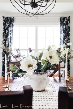 a dining room table with white flowers in a vase on the centerpiece and candles