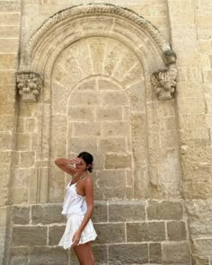 a woman leaning against a stone wall with her hand on her head and looking at the camera