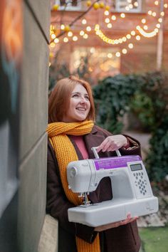 a woman standing next to a wall with a sewing machine in her hand and smiling