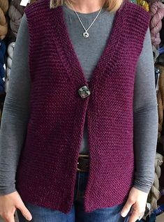 a woman standing in front of a pile of yarns wearing a purple sweater vest