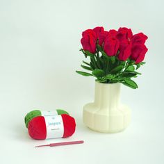 a white vase filled with red roses next to a crochet ball and needles