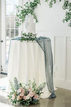 a white wedding cake with greenery on top and flowers around the bottom, sitting on a table