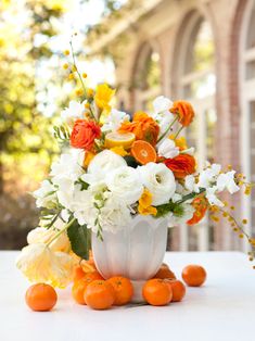 an arrangement of oranges and white flowers in a vase on a table with greenery