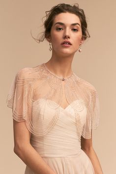 a woman wearing a white dress with sheer sleeves and beading on the shoulders, in front of a beige background