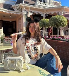 a woman sitting at an outdoor table with a mickey mouse t - shirt on and holding a toothbrush