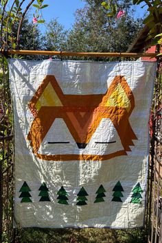 an orange and white quilt hanging from a metal fence with trees on it's sides