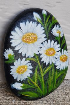 a painted rock with white daisies and green leaves on the outside, next to a wall