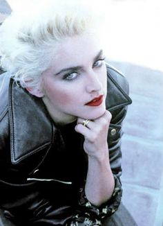 a woman with blond hair wearing a black leather jacket and red lipstick sitting on a bench