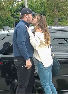 a man and woman kissing in front of a black car with trees in the background