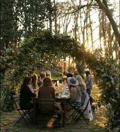 a group of people sitting around a table in the middle of a forest at sunset