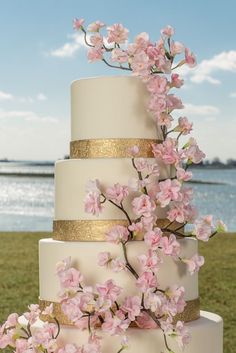 a white and gold wedding cake with pink flowers on the top tier, in front of water