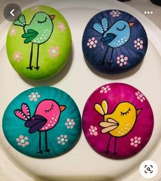 four painted rocks sitting on top of a white plate with flowers and birds in different colors