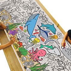 two children sitting at a table drawing on a large sheet of paper with animals and fish