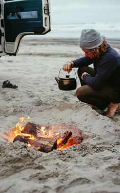 a man sitting in the sand next to a campfire with a bucket on it