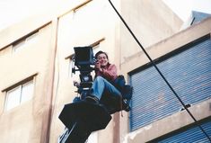 a man sitting on top of a camera next to a tall building with blue shutters