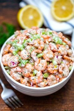 a white bowl filled with shrimp salad next to a fork and lemon wedges on the side