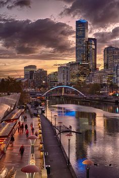 people are walking along the water in front of some tall buildings at sunset or dawn