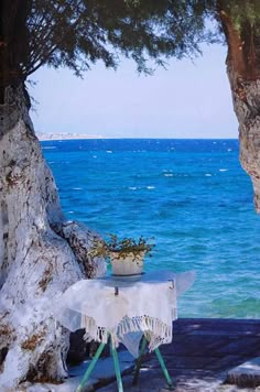 a table with a white cloth on it sitting next to the ocean and some trees