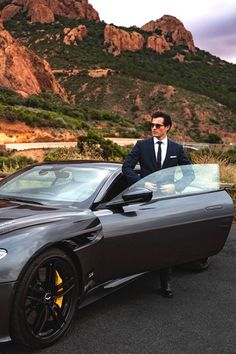 a man in a suit standing next to a sports car with mountains in the background