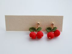 two red cherries with leaves on them are attached to gold earwires, sitting on a white surface