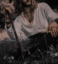 a man with long hair sitting on the ground holding a stick and wearing a white shirt