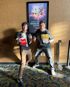 two men standing in front of a movie poster holding up drinks and popcorn boxes while posing for the camera