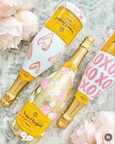 two bottles of champagne next to pink flowers on a gray surface with white and gold lettering